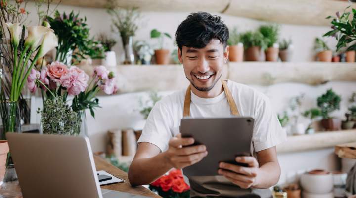 A person in a plant shop is using a tablet and laptop at a wooden table, surrounded by shelves of potted plants and flowers.